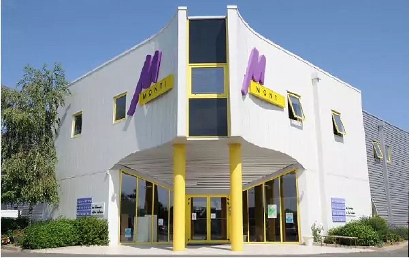 Façade d'un bâtiment moderne blanc aux accents jaunes, orné du logo violet et de l'enseigne jaune de l'entreprise, avec une entrée sous un porche à colonnes jaunes.