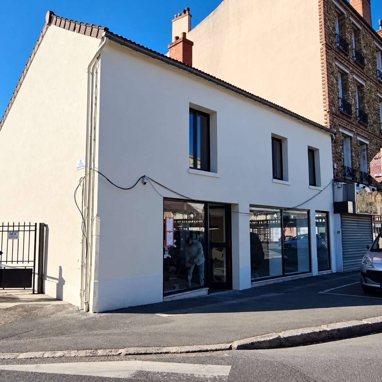 Façade blanche d'un bâtiment d'angle avec de grandes vitrines au rez-de-chaussée, donnant sur une rue sous un ciel bleu.