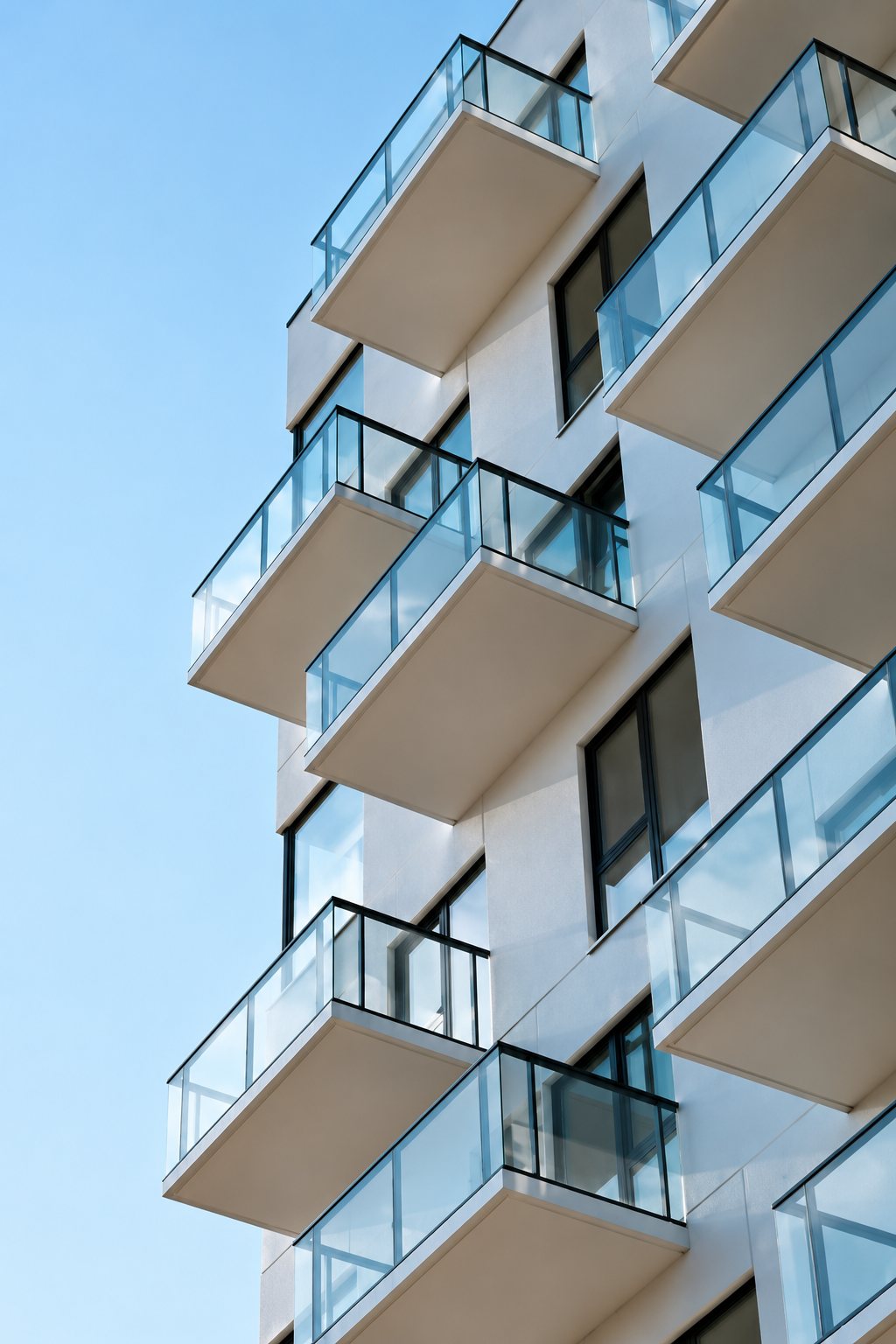 Vue en contre-plongée d'un immeuble moderne blanc dont les balcons sont équipés de garde-corps en verre, sous un ciel bleu.