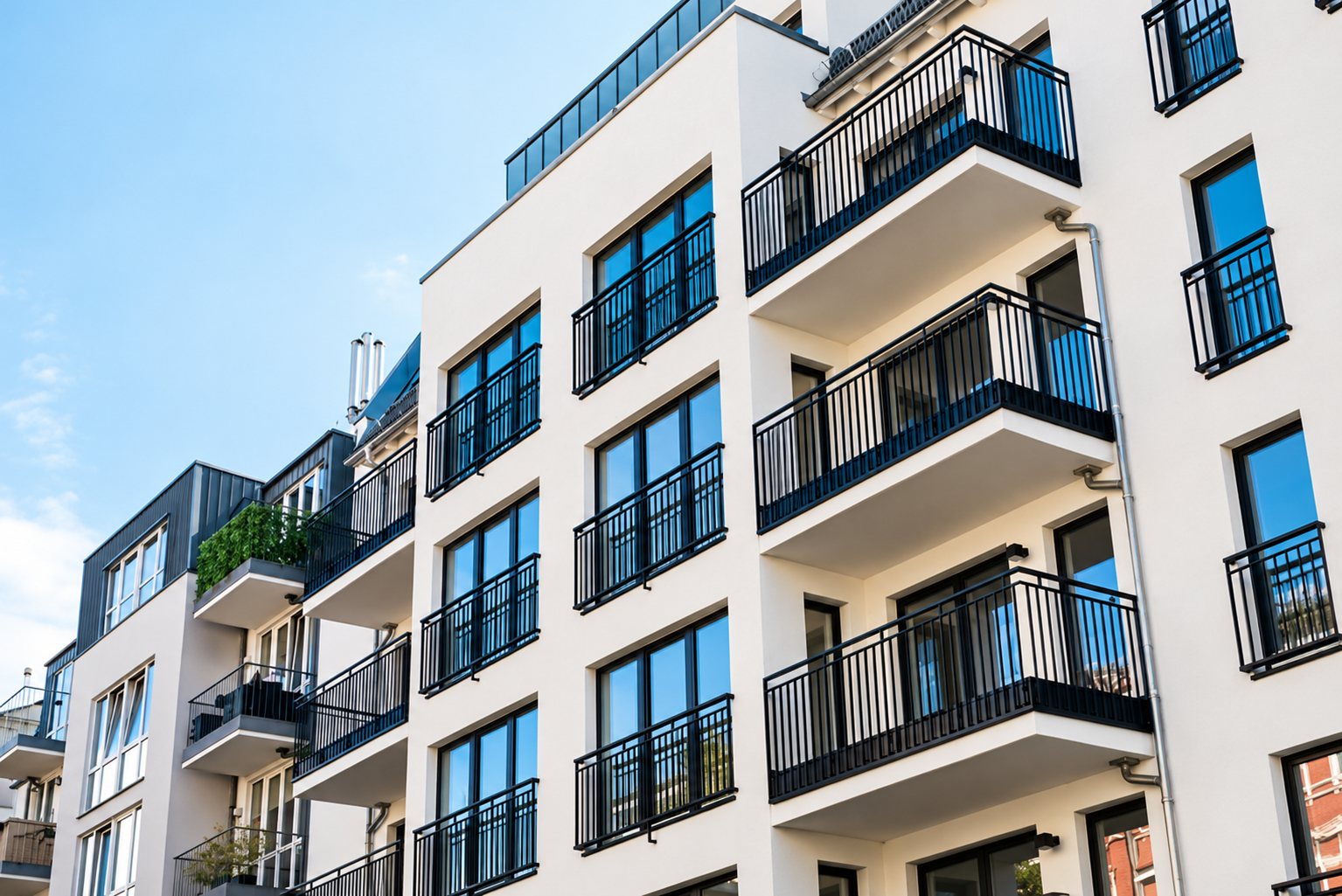 Vue en contre-plongée d'un immeuble d'habitation moderne aux façades blanches, avec grandes fenêtres et balcons à garde-corps métalliques noirs sous un ciel bleu.
