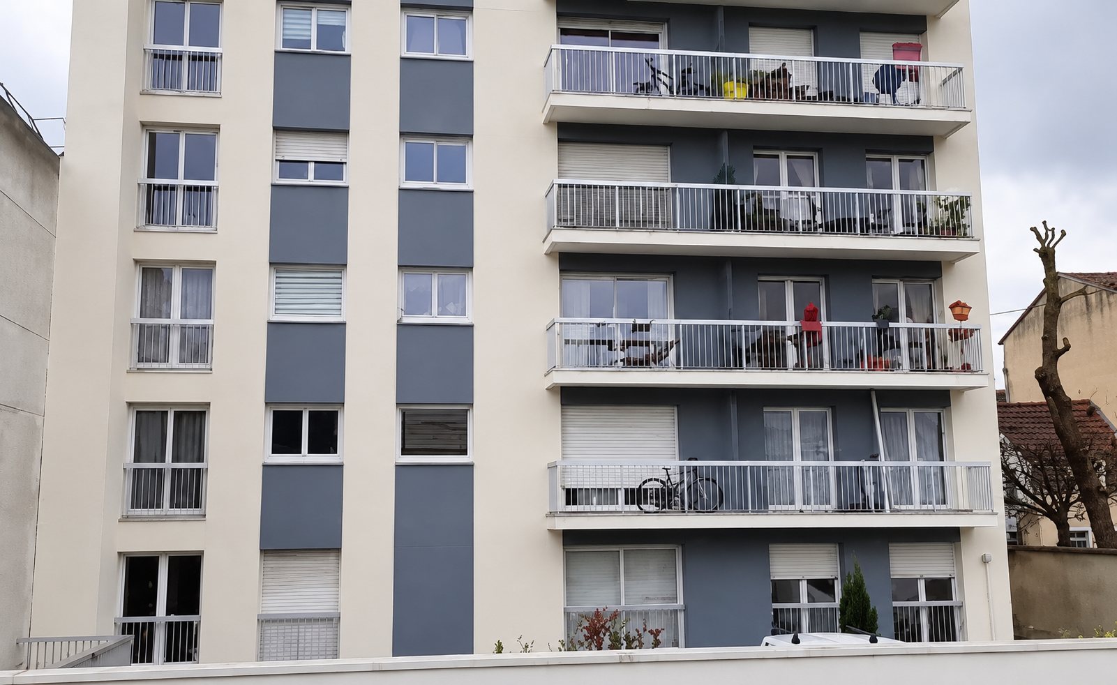 Façade d'un immeuble d'habitation récent aux murs clairs et panneaux gris-bleu, avec plusieurs balcons à garde-corps métalliques.
