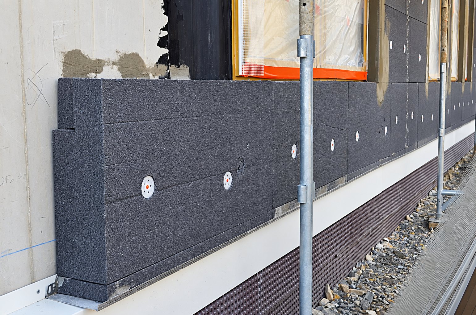 Chantier d'isolation thermique par l'extérieur où des panneaux de polystyrène gris sont fixés par des chevilles blanches sur le mur d'un bâtiment, avec un échafaudage au premier plan.
