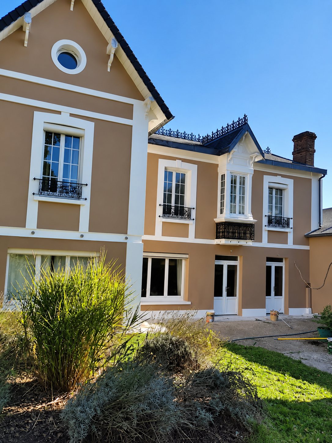 Façade d'une maison ancienne rénovée, peinte en beige avec des encadrements de fenêtres blancs et des garde-corps en fer forgé, sous un ciel bleu.