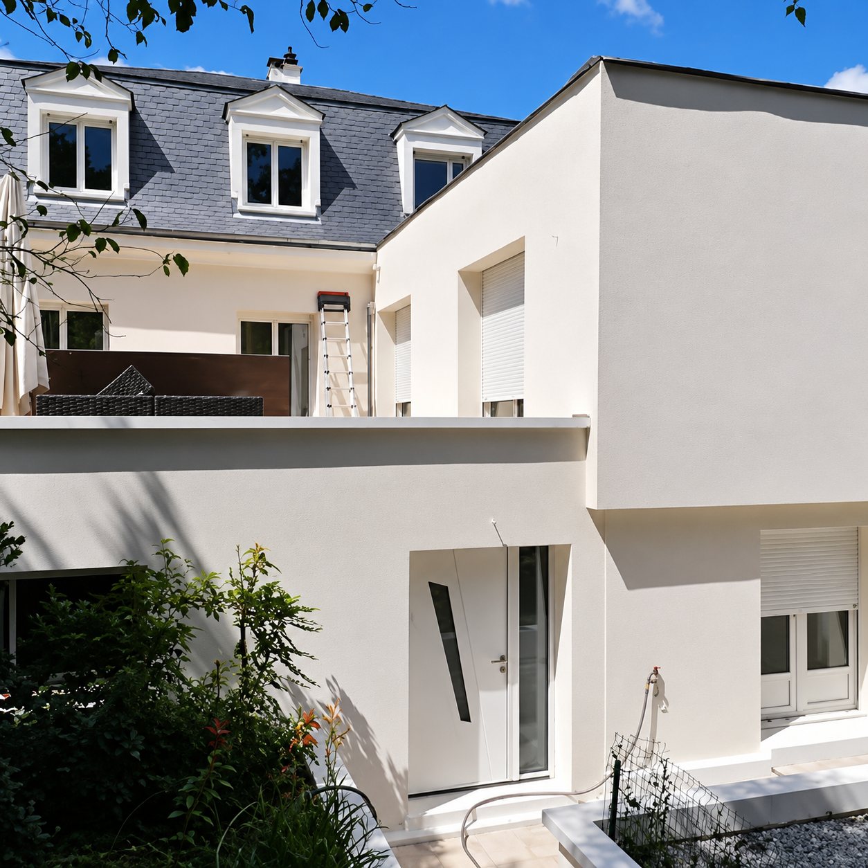 Façade d'une maison rénovée avec enduit clair, lucarnes sur un toit en ardoise, extension moderne et échelle appuyée contre le mur.