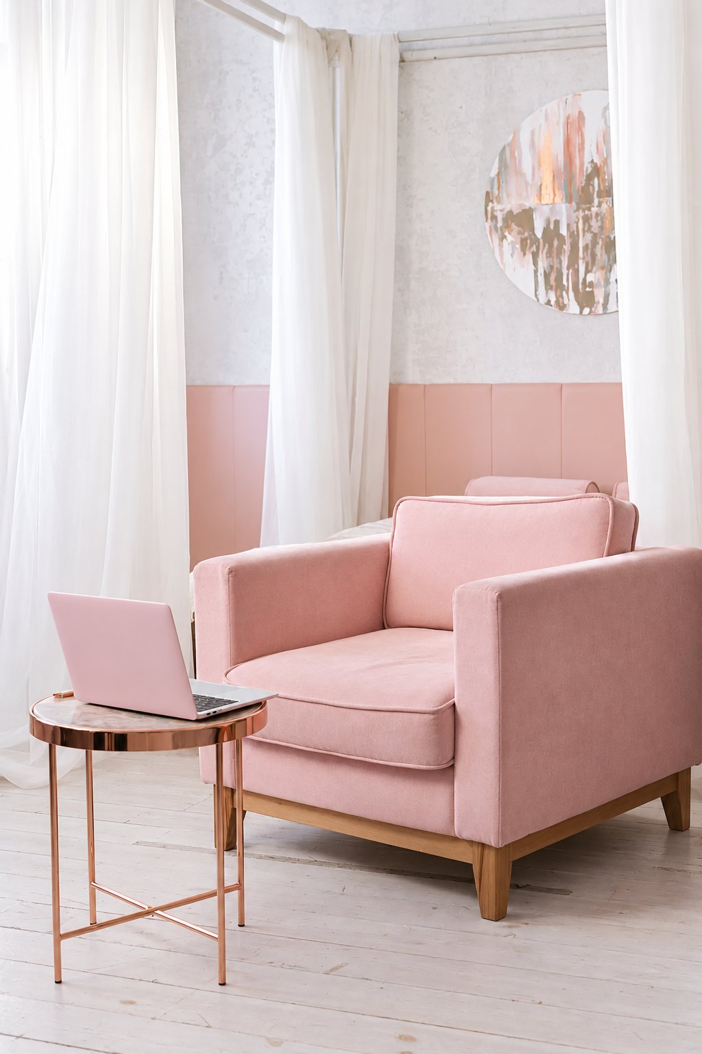 Un fauteuil rose en velours aux pieds en bois, à côté d'une petite table ronde cuivrée où est posé un ordinateur portable rose, devant des voilages blancs.