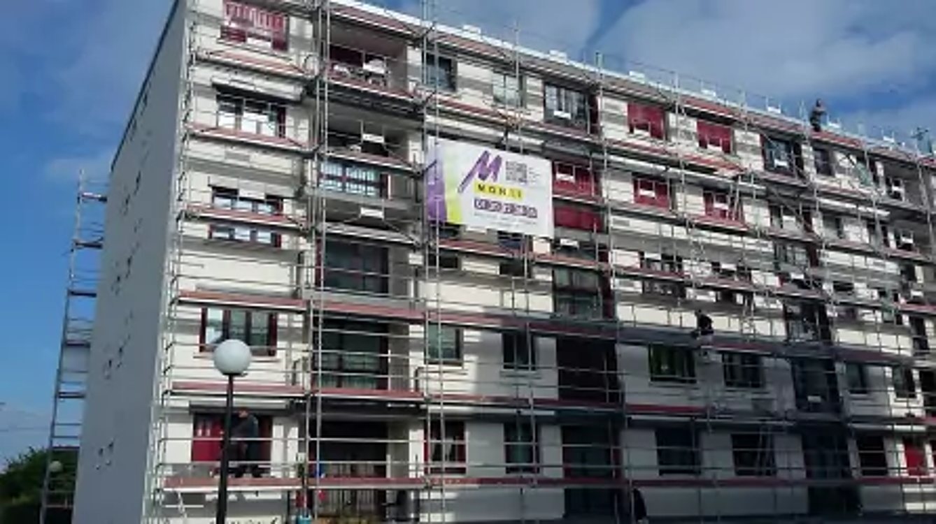 Façade d'un immeuble de plusieurs étages entièrement couverte d'échafaudages, avec une grande bannière publicitaire accrochée, sous un ciel bleu.