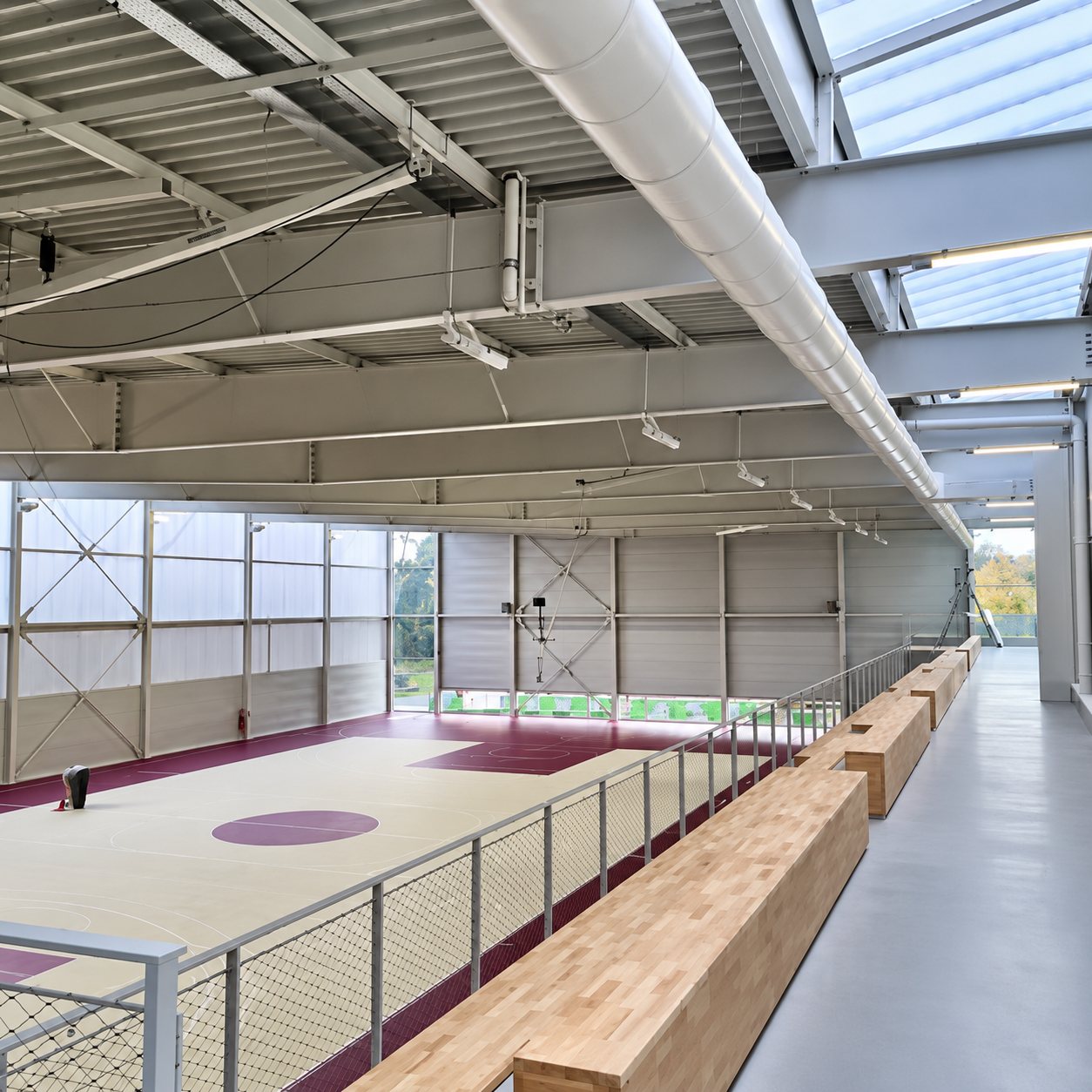 Intérieur d'un gymnase récemment construit avec charpente métallique, gaine blanche au plafond, terrain au sol clair et bancs en bois le long d'une coursive.