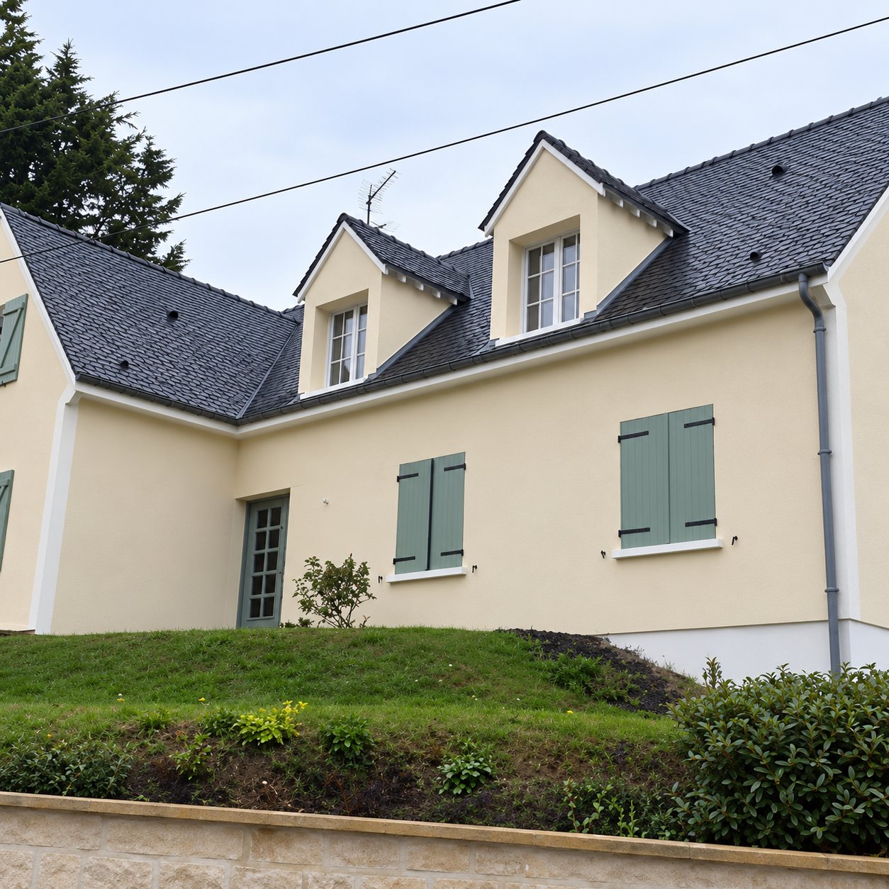 Maison à façade beige avec lucarnes, toit en ardoise sombre et volets vert-gris, posée sur un talus herbeux.