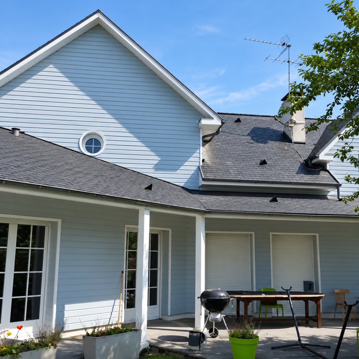 Façade arrière d'une maison à clin bleu clair avec toiture en ardoise, une terrasse couverte, un barbecue et une table en bois.