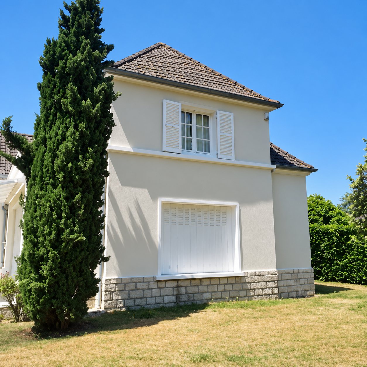 Maison à deux niveaux à la façade enduite beige clair, avec volets blancs, soubassement en pierre et grand cyprès sur une pelouse sèche sous un ciel bleu.