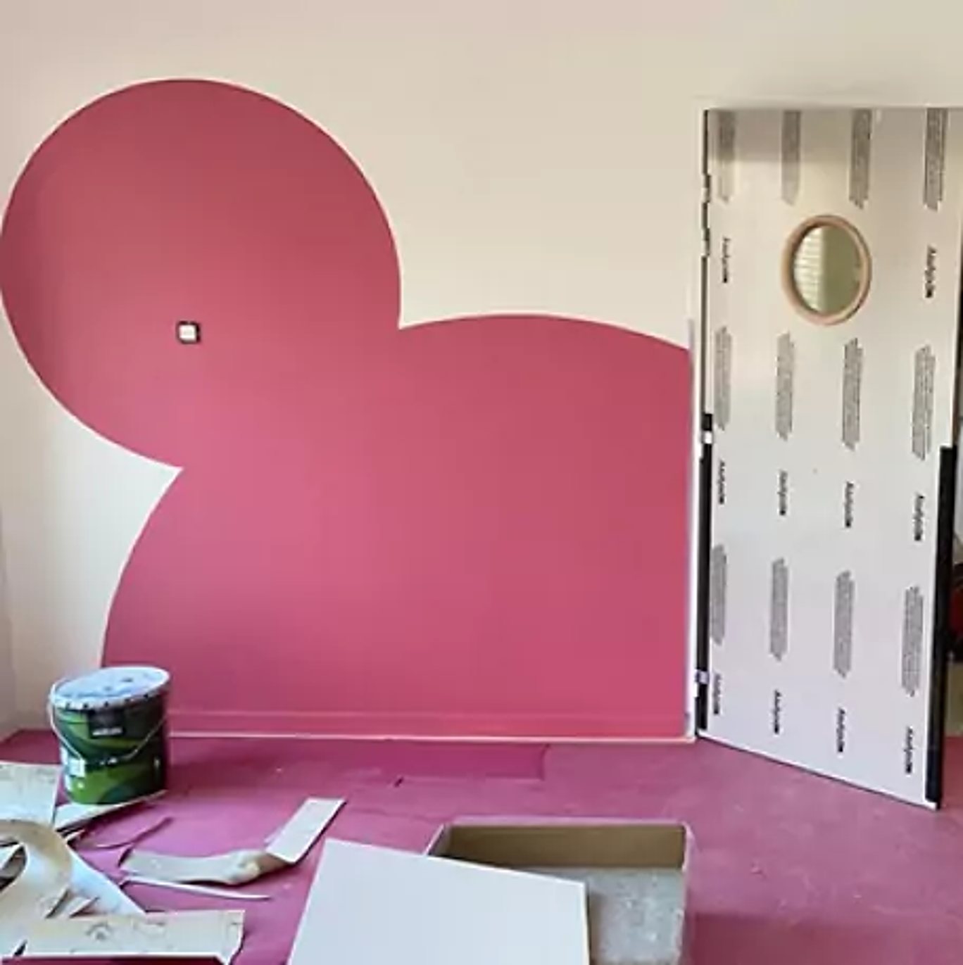 Dans une pièce en travaux, un mur blanc est décoré d'un grand motif rose arrondi en forme de souris, à côté d'une porte protégée par un film et d'un pot de peinture posé sur le sol rose.