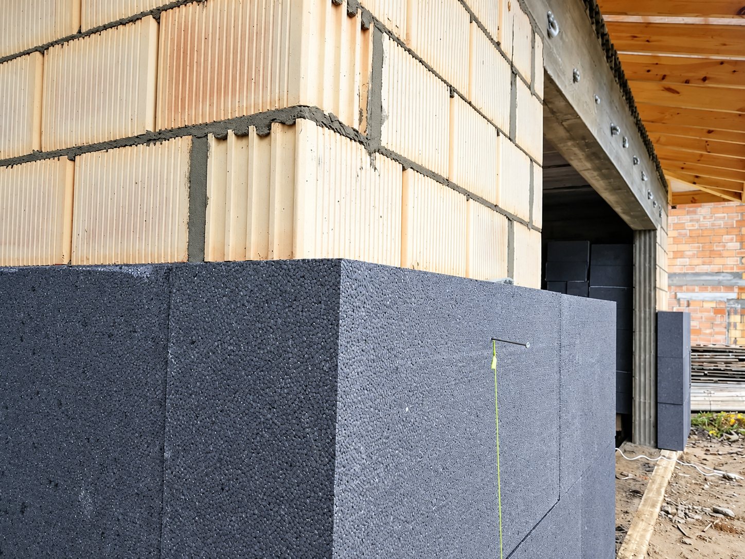 Sur un chantier de construction, des panneaux d'isolation en polystyrène gris foncé sont fixés au bas d'un mur en briques alvéolées beige.