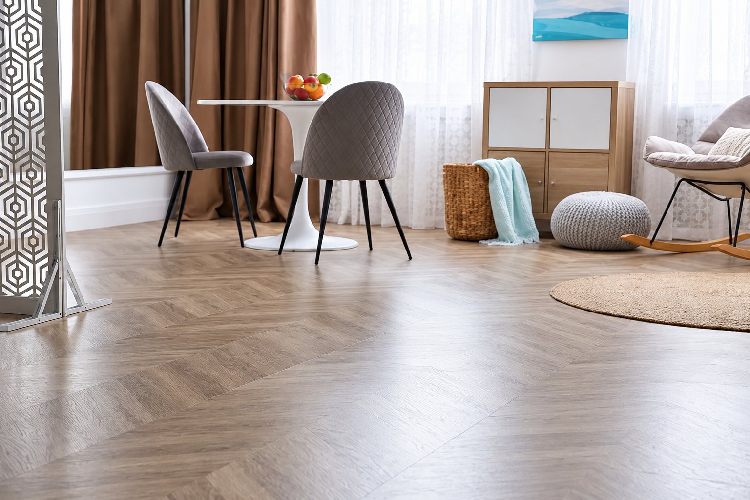 Un salon lumineux avec un sol en parquet à chevrons gris, une table ronde blanche, deux chaises en velours gris, un pouf tricoté et un fauteuil à bascule.