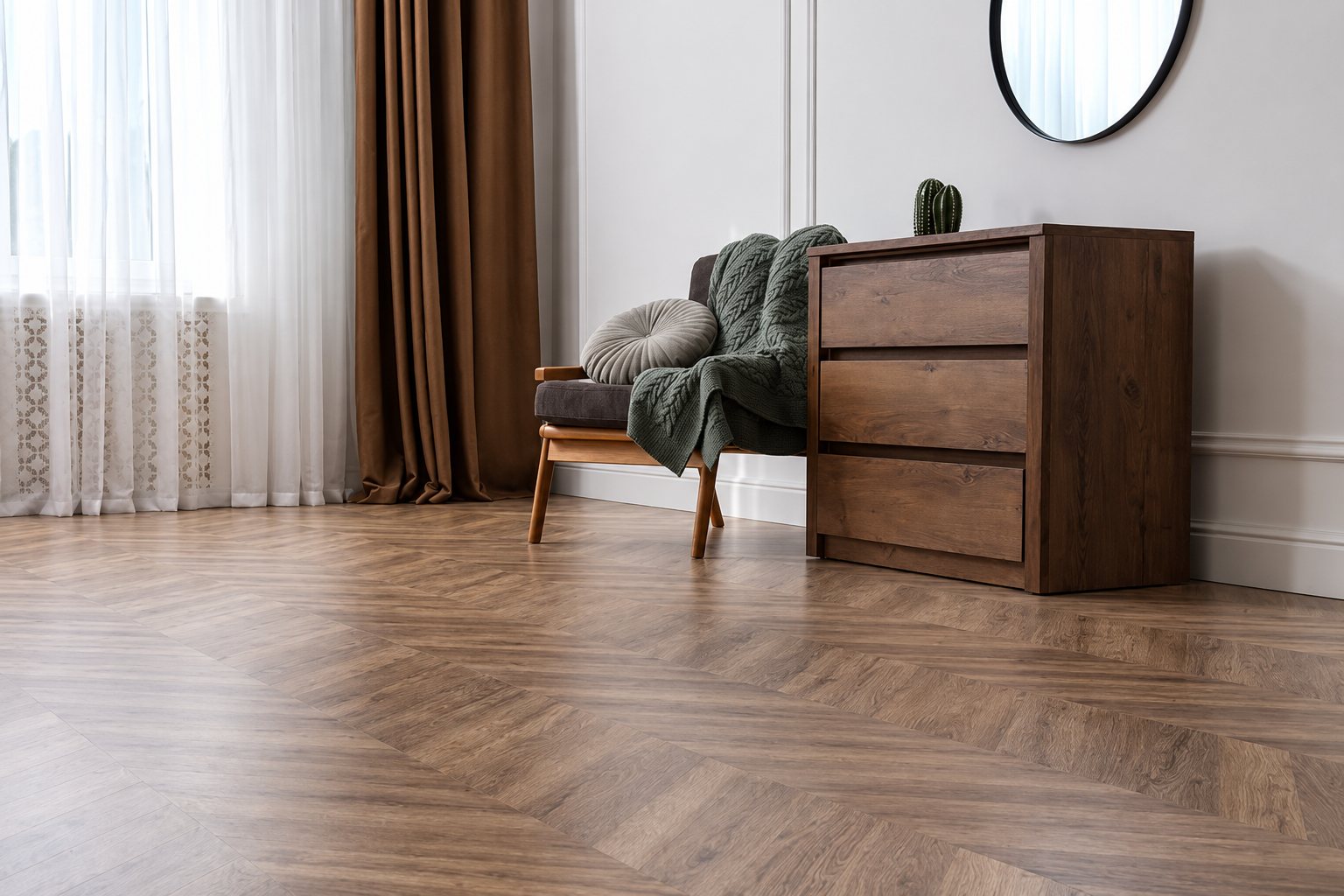 Un sol en parquet à chevrons gris-brun occupe un salon lumineux meublé d'une commode en bois, d'un fauteuil avec plaid vert, de rideaux marron et d'un miroir rond.