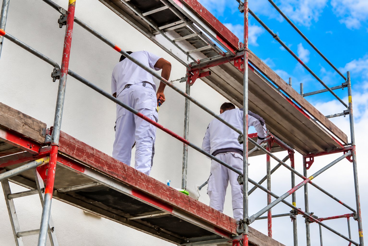 Deux ouvriers en tenue blanche travaillent sur un échafaudage pour peindre ou enduire la façade d'un bâtiment sous un ciel bleu.