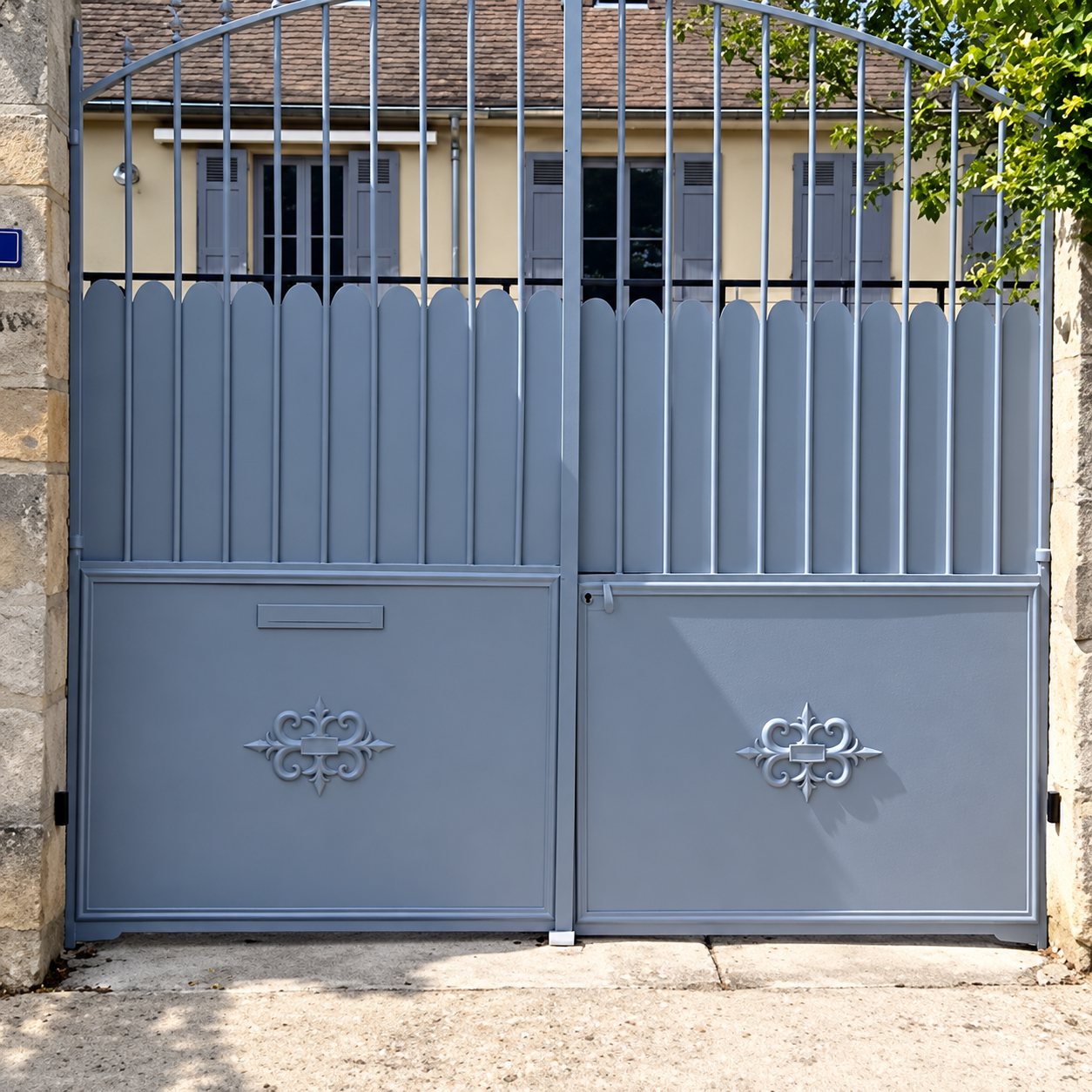 Un grand portail à deux battants en métal gris, orné de motifs en fer forgé, ferme l'entrée d'une propriété devant une maison.