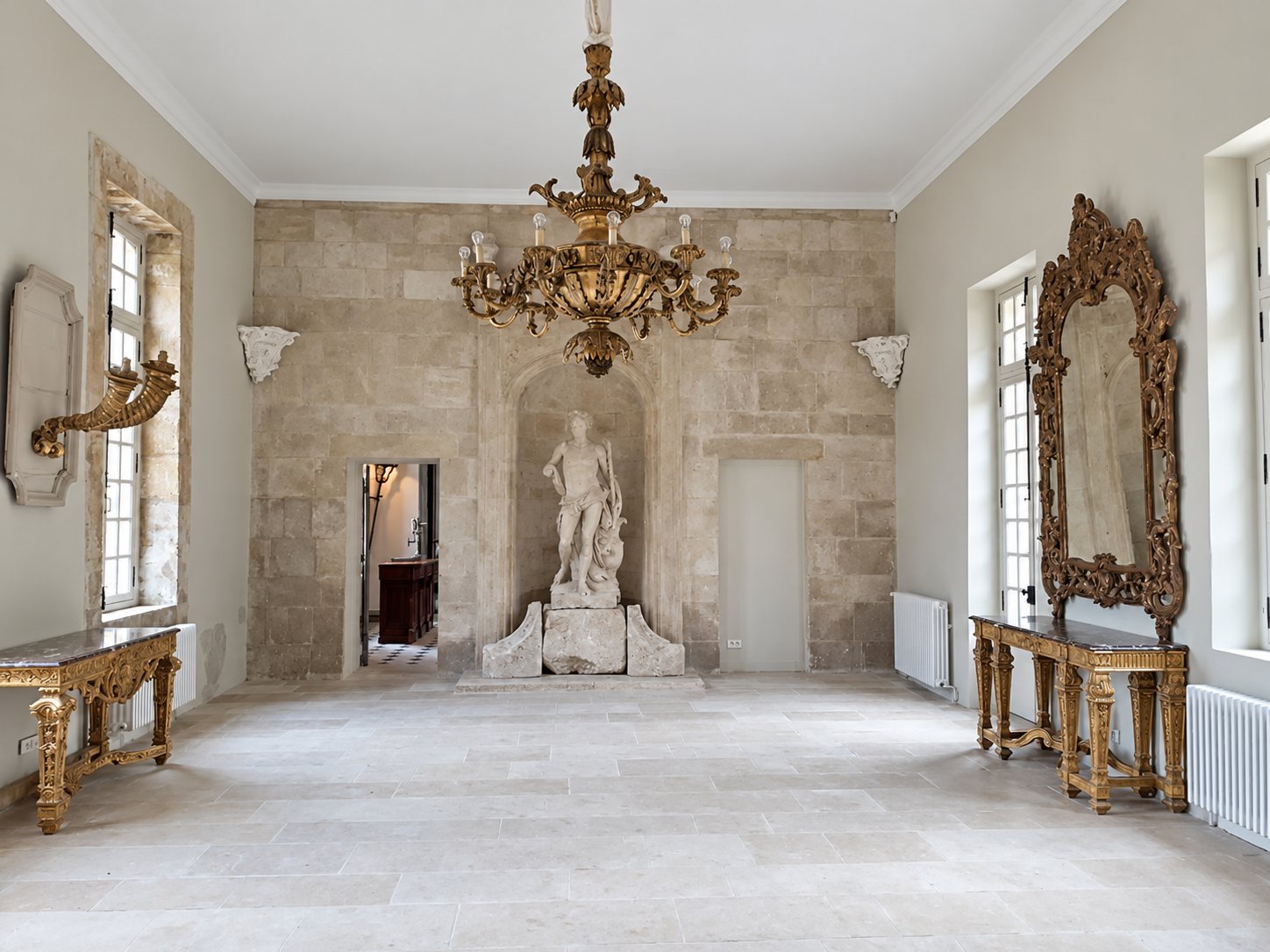 Une grande salle aux murs de pierre claire, ornée d'un lustre doré, d'une statue blanche dans une niche, de deux consoles dorées et d'un grand miroir sculpté.