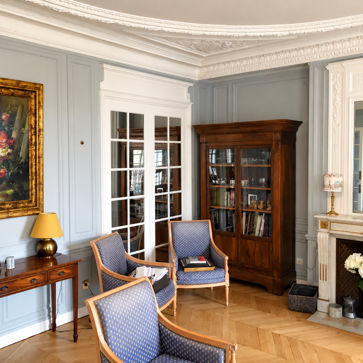 Salon haussmannien aux boiseries claires, avec parquet en chevrons, bibliothèque ancienne en bois, fauteuils bleus, cheminée et tableau doré.