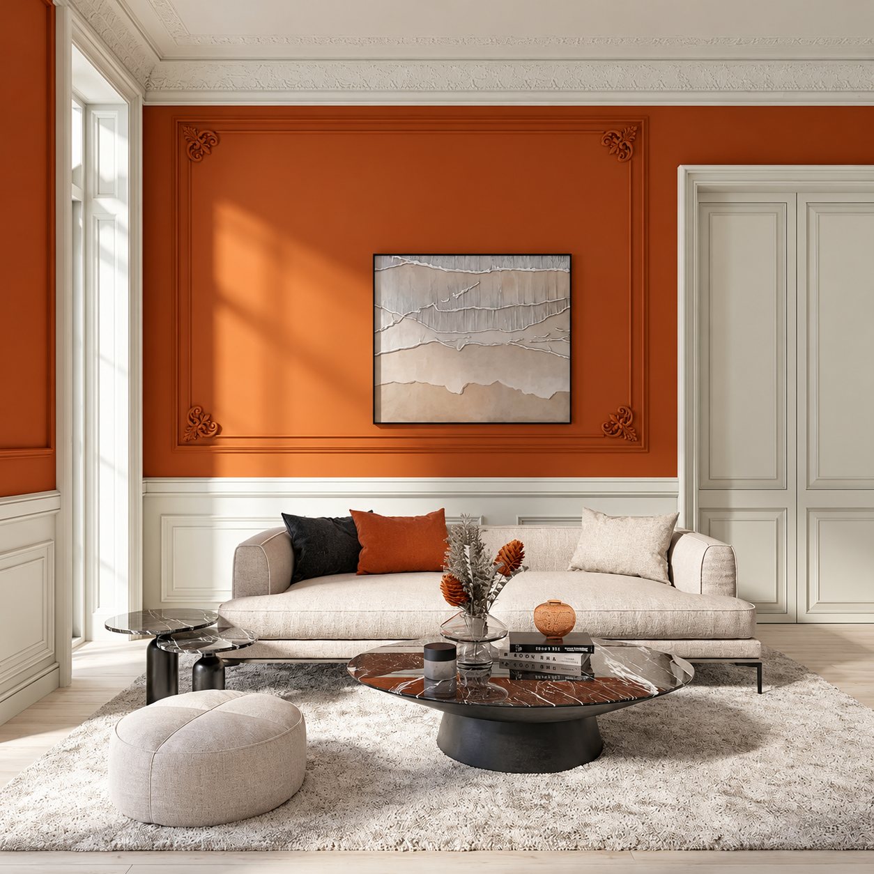 Salon lumineux aux murs terracotta et moulures blanches, avec un canapé beige, une table basse ronde en marbre, un pouf et un tableau abstrait.