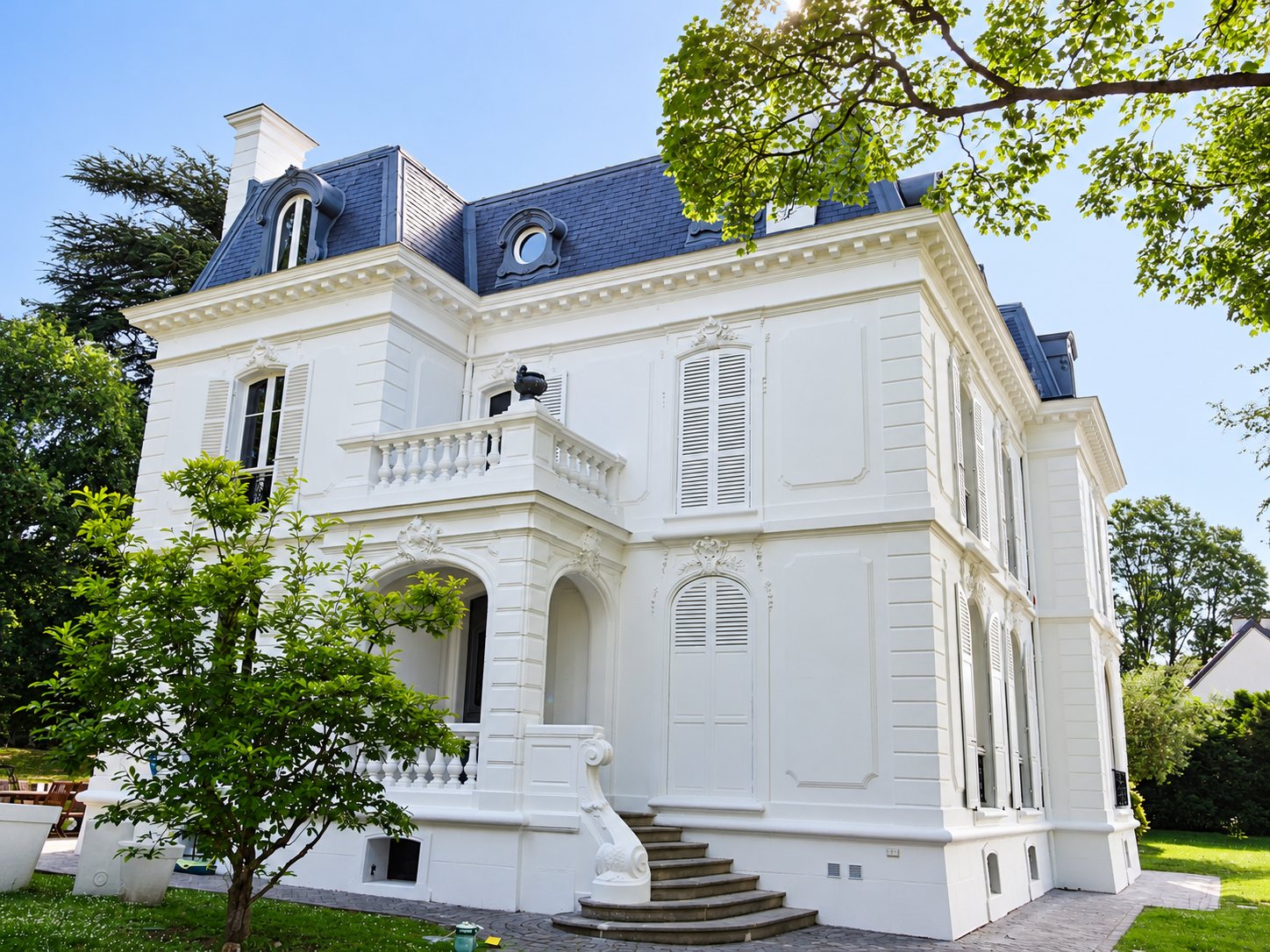 Façade d'une élégante villa blanche aux volets clairs, au toit mansardé en ardoise et à l'escalier en pierre, entourée d'arbres et de pelouse.